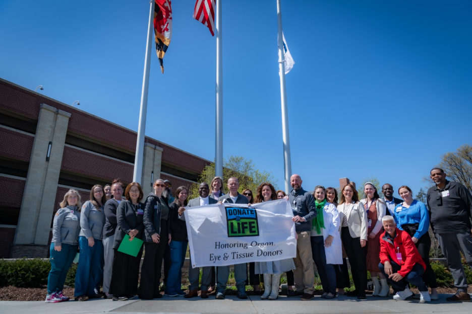 Frederick Health and Infinite legacy members gather in front of a flag pole, holding up a flag that says "Donate Life, Honoring Organ, Eye, and Tissue Donors." Approximately 20 people in the photo.