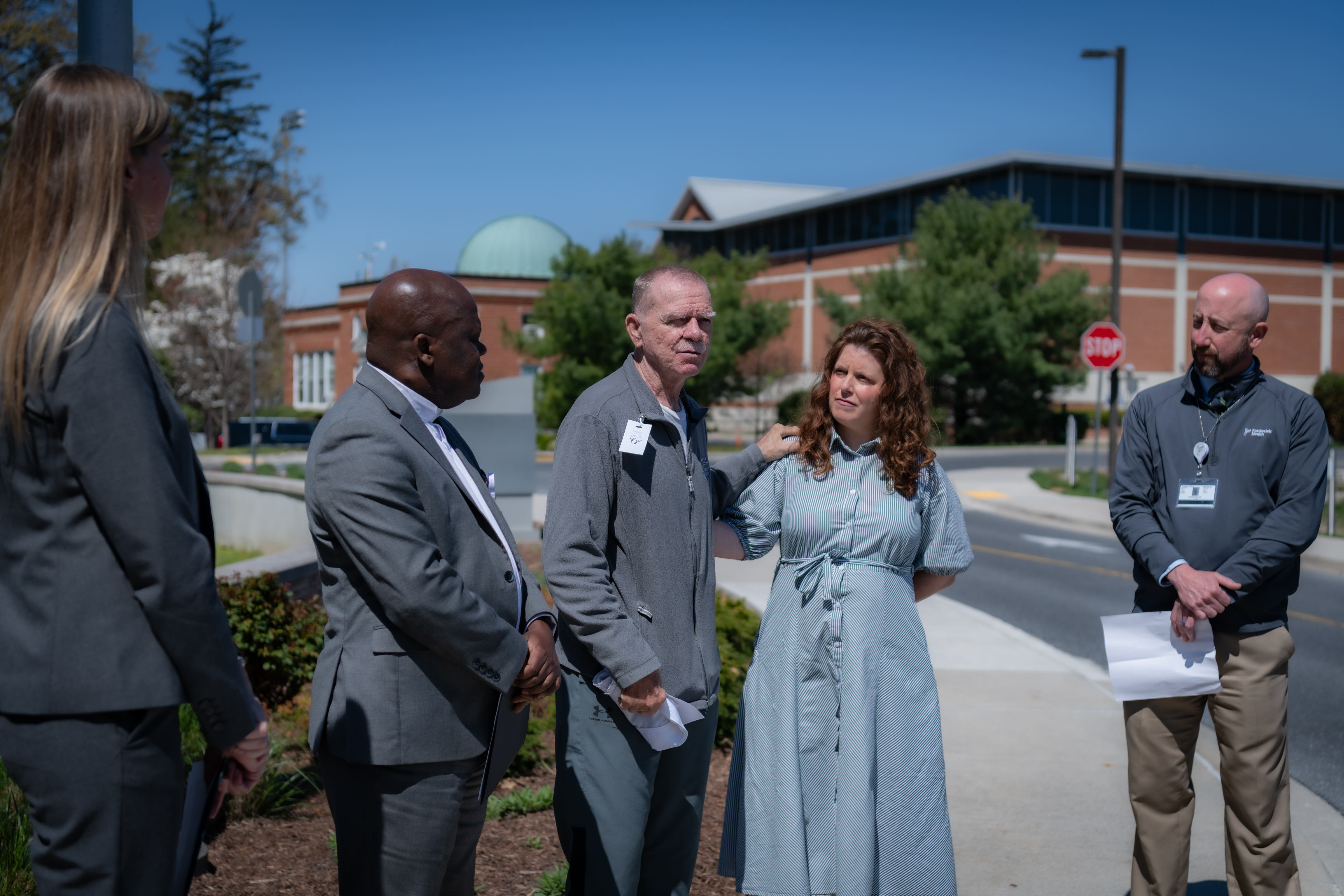 Five people stand on a clear day in front of a brick building, three men and two women. One of the man discusses his experience as a donor recipient