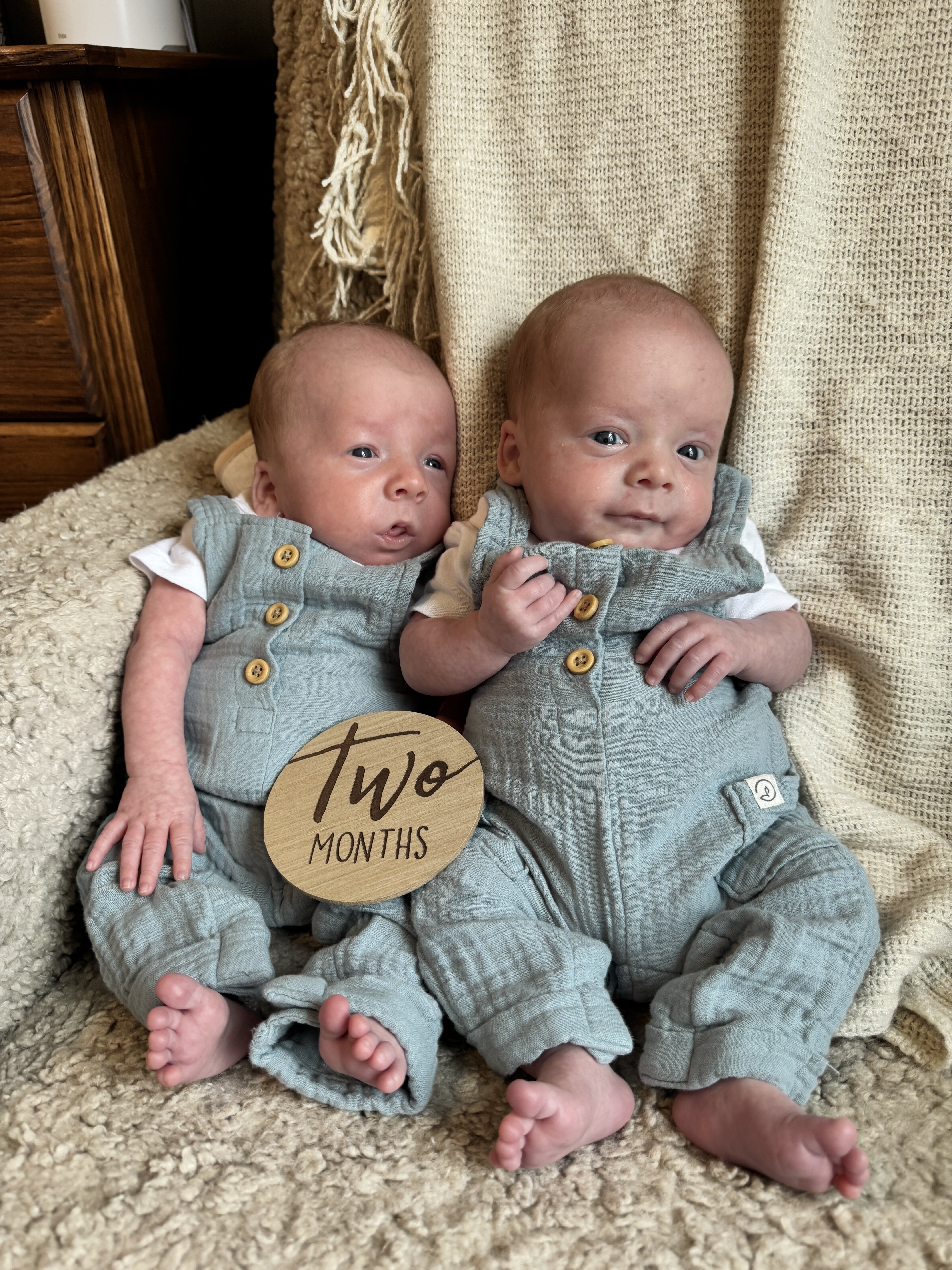 Two babies in blue clothes sitting on a couch with a "two months" wooden plaque