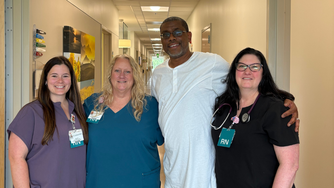 Three women in scrubs with nurse badges stand with arms wrapped around one another with one taller man, a former cancer patient. All are smiling at the camera.