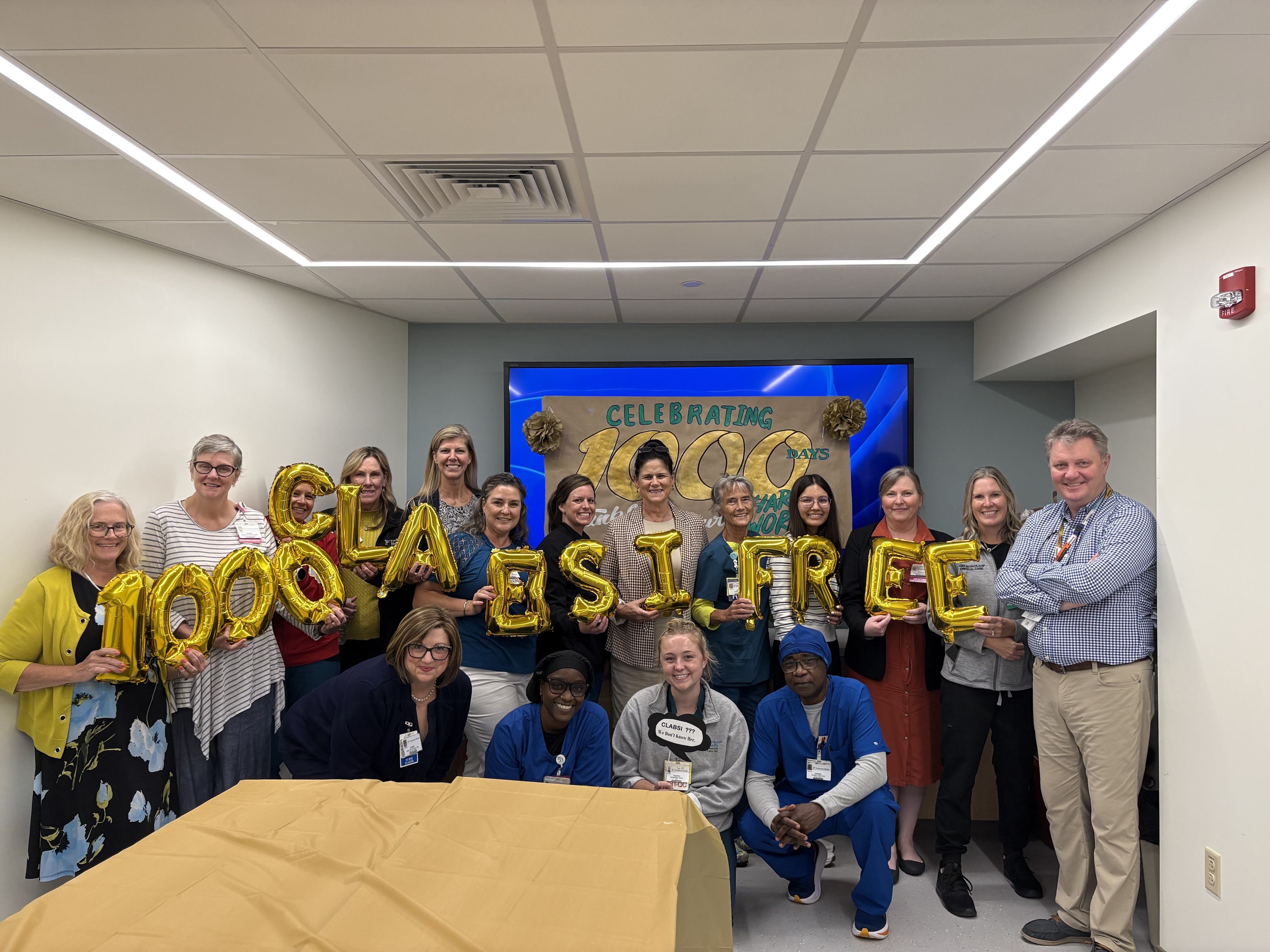 A group of smiling people stand in a conference room carrying foil balloons that read "1,000 CLABSI Free"